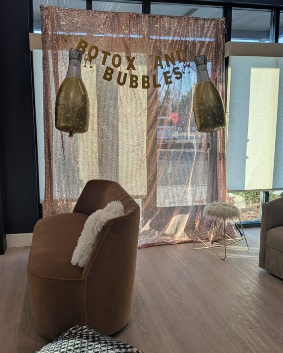 Three smiling women pose beneath the ‘Botox and Bubbles’ sign against a rose-gold sequin backdrop.