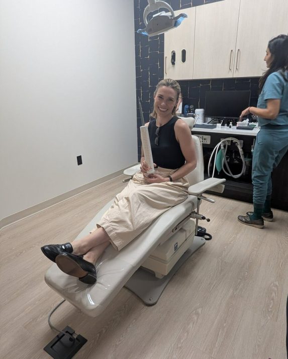 A smiling woman sits in a treatment chair holding an oversized syringe prop while a clinician prepares supplies.