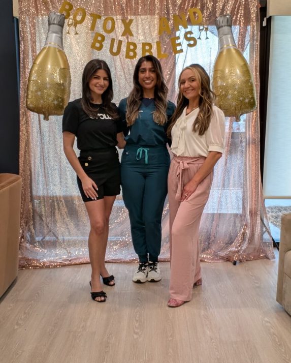 Three smiling women pose beneath the ‘Botox and Bubbles’ sign against a rose-gold sequin backdrop.