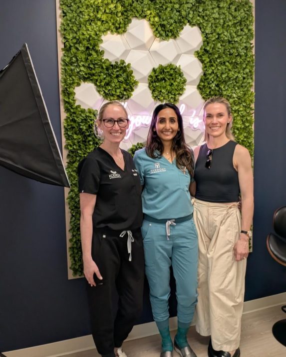 Three smiling women pose together in front of a green-and-white floral feature wall.